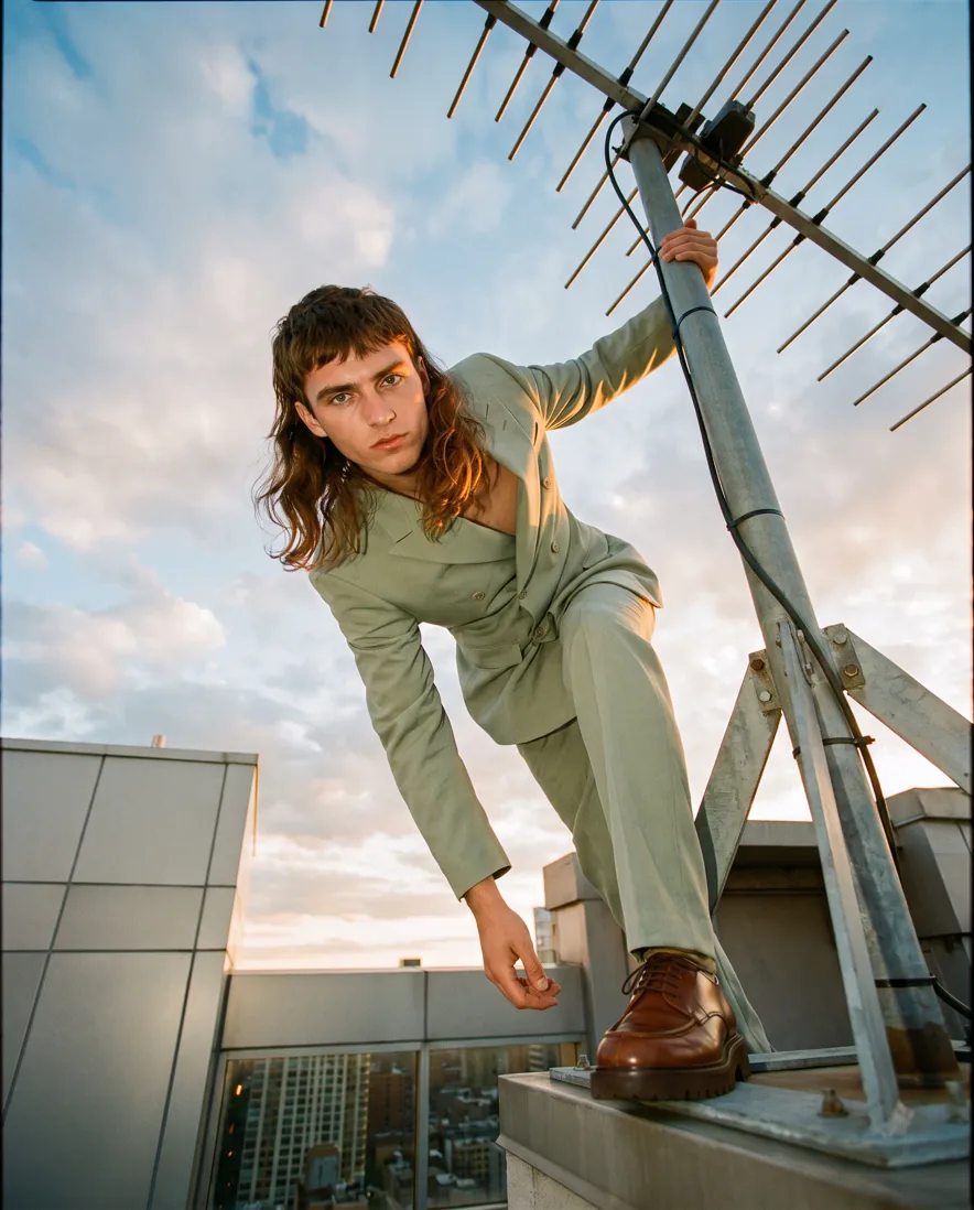 Model in a sage suit climbing a rooftop antenna at sunset